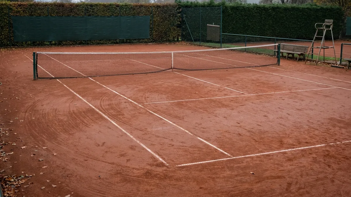 Leerer Tennisplatz mit rotem Sandbelag, gespanntem Netz und Schiedsrichterstuhl unter bedecktem Himmel