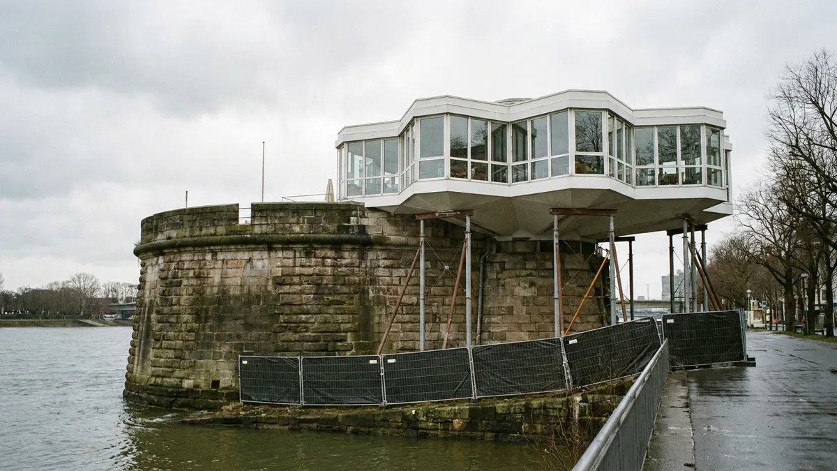 Verglaster Aufbau auf einem runden Steinsockel am Flussufer, gestützt von Stahlstreben und umstellt von Bauzäunen