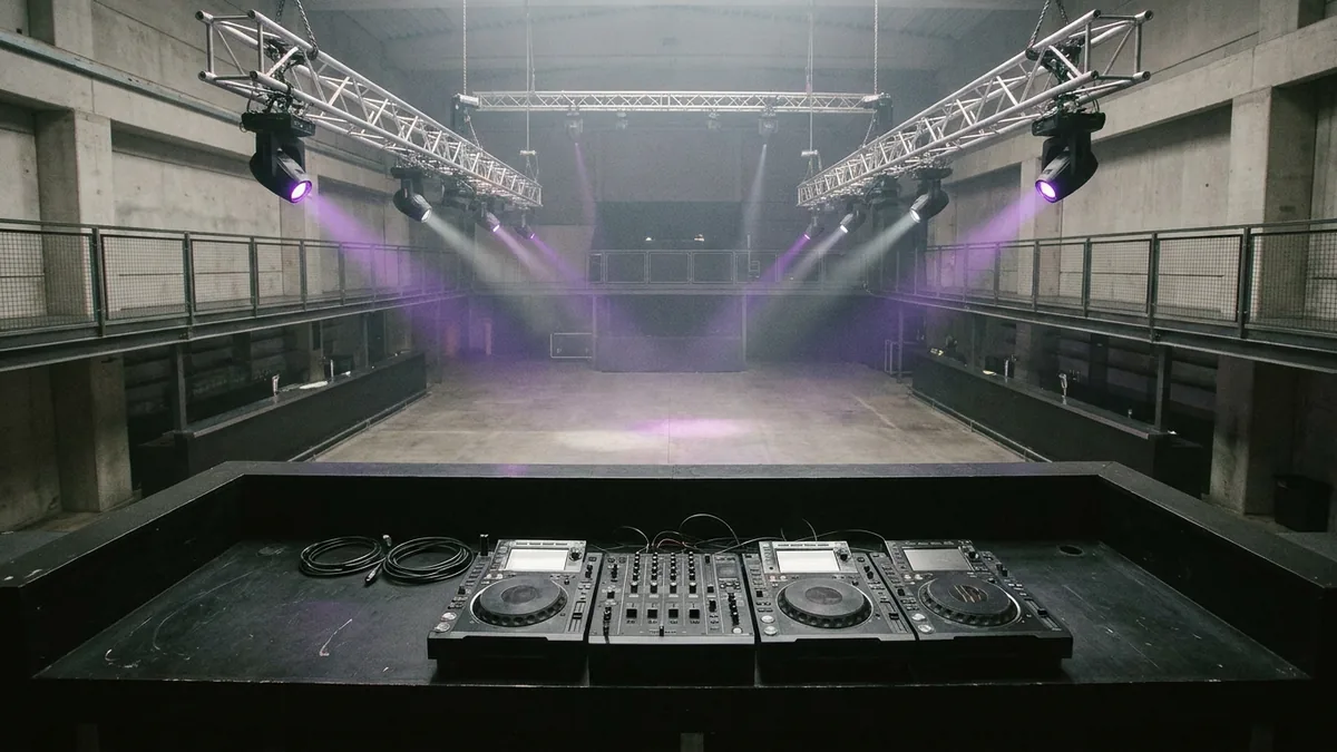 Blick über ein DJ-Pult mit Playern und Mischpult in einen leeren Hallensaal, violette Lichtkegel im Nebel