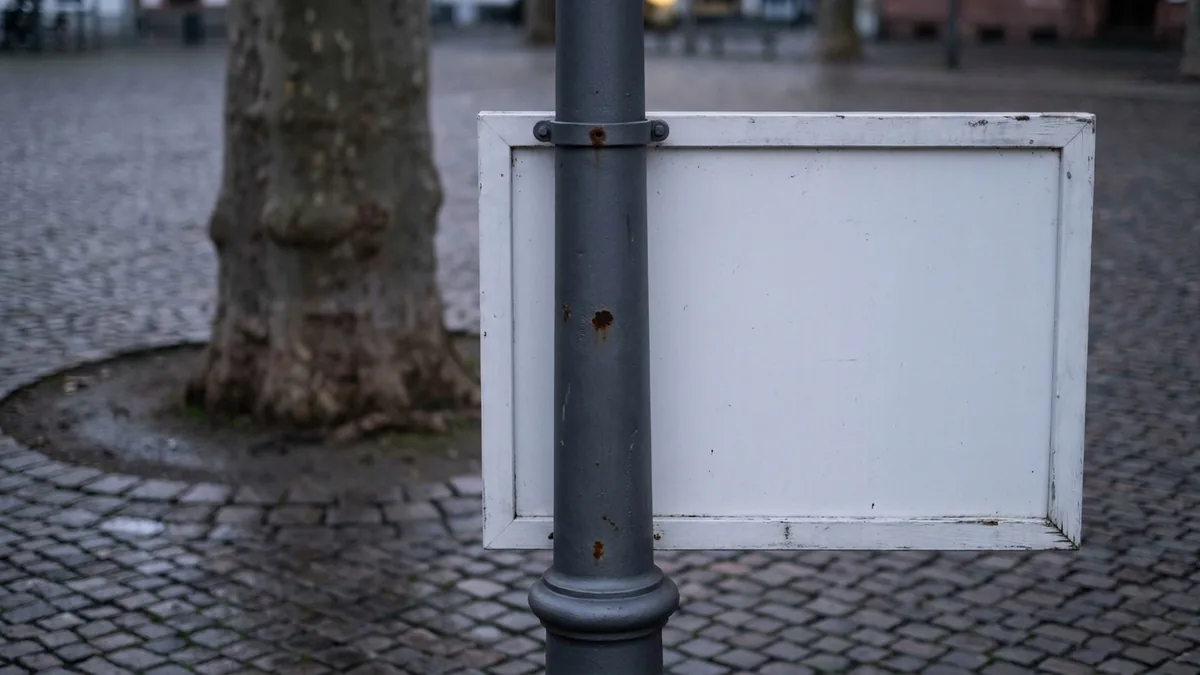 Leere weiße Tafel an einem grauen Metallmast auf einem gepflasterten Platz, dahinter ein Baumstamm