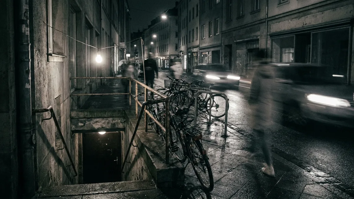 Kellerabgang an einer nächtlichen Straße, davor Fahrräder an einem Geländer und eine Glühbirne an einem Kabel