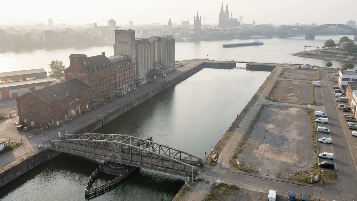 Luftaufnahme eines Hafenbeckens mit Drehbrücke, alten Mühlengebäuden und Silos, im Hintergrund Rhein und Kölner Dom