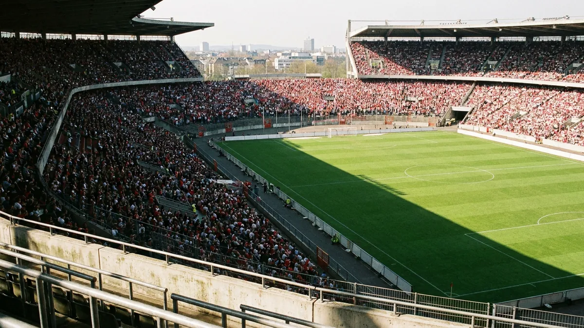 Blick von der Tribüne in ein gut gefülltes Fußballstadion, viele Zuschauer in Rot und Weiß, sonniger Nachmittag