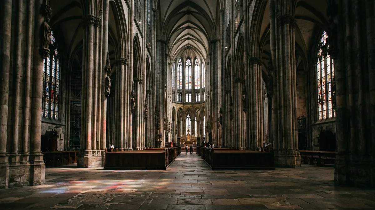 Mittelschiff einer gotischen Kathedrale, farbiges Licht fällt aus den Fenstern auf den Steinboden.