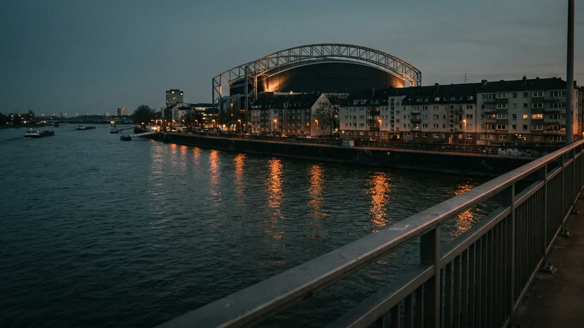 Blick von einer Brücke über einen Fluss in der Dämmerung, am Ufer Wohnhäuser, dahinter eine Arena mit beleuchtetem Bogendach