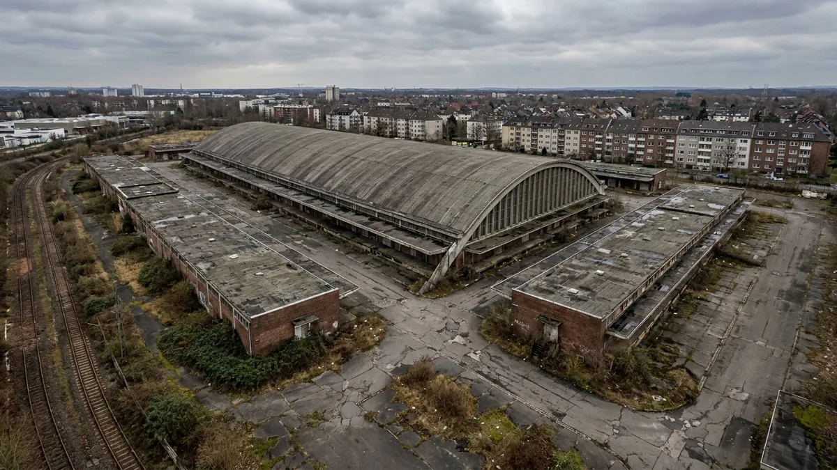 Luftaufnahme einer verlassenen Halle mit langem Tonnendach, rissigem Asphalt und Gleisen, dahinter Wohnblocks