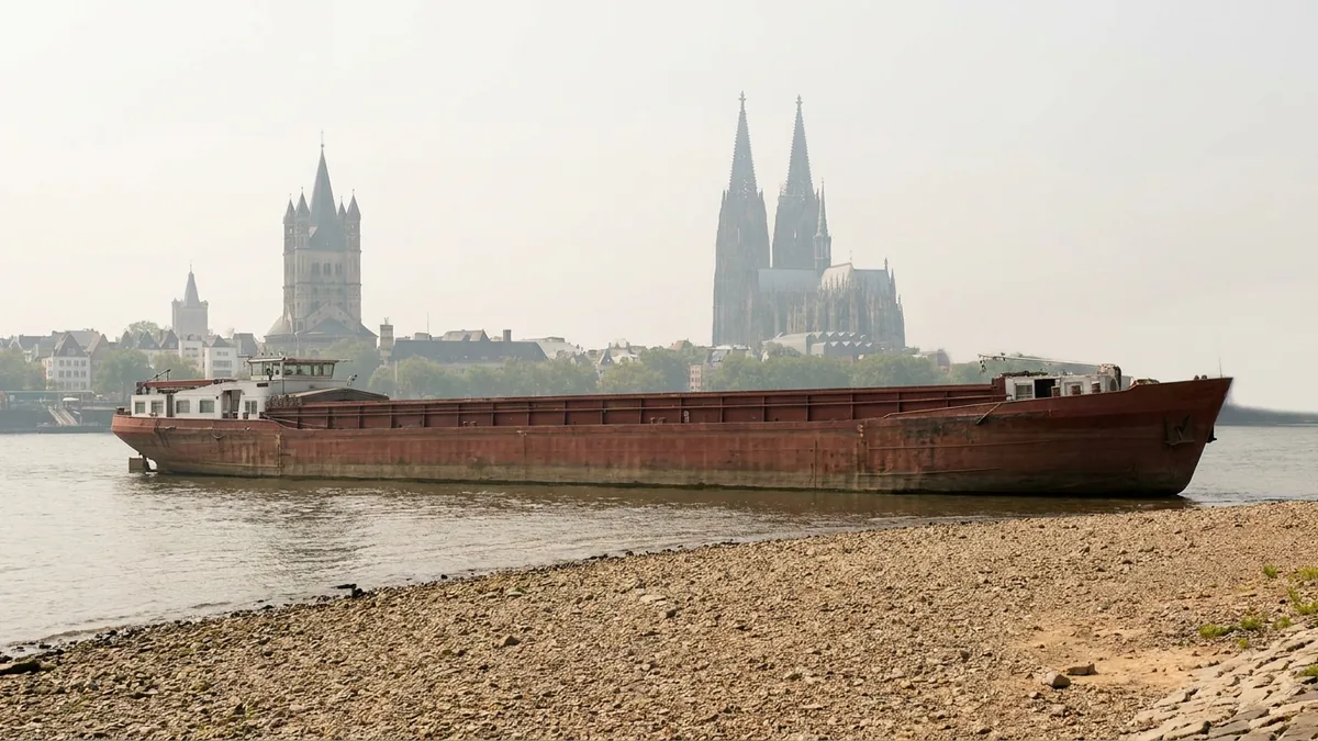 Frachtschiff ragt hoch aus dem Rhein, vorn eine breite Kiesbank, dahinter Groß St. Martin und der Kölner Dom im Dunst