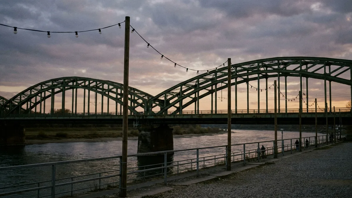 Stählerne Bogenbrücke über einem Fluss in der Abenddämmerung, davor Lichterketten zwischen Holzmasten